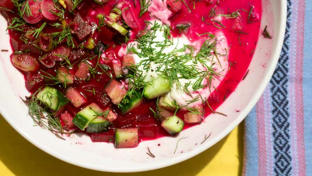Close-up, top-down photo of a bowl of cold borsch with cucumbers, scallions, dill, and sour cream. About half the white bowl is visible. You can see a small sliver of yellow tablecloth at the bottom of the frame, and a light blue and pink striped napkin to the right.   